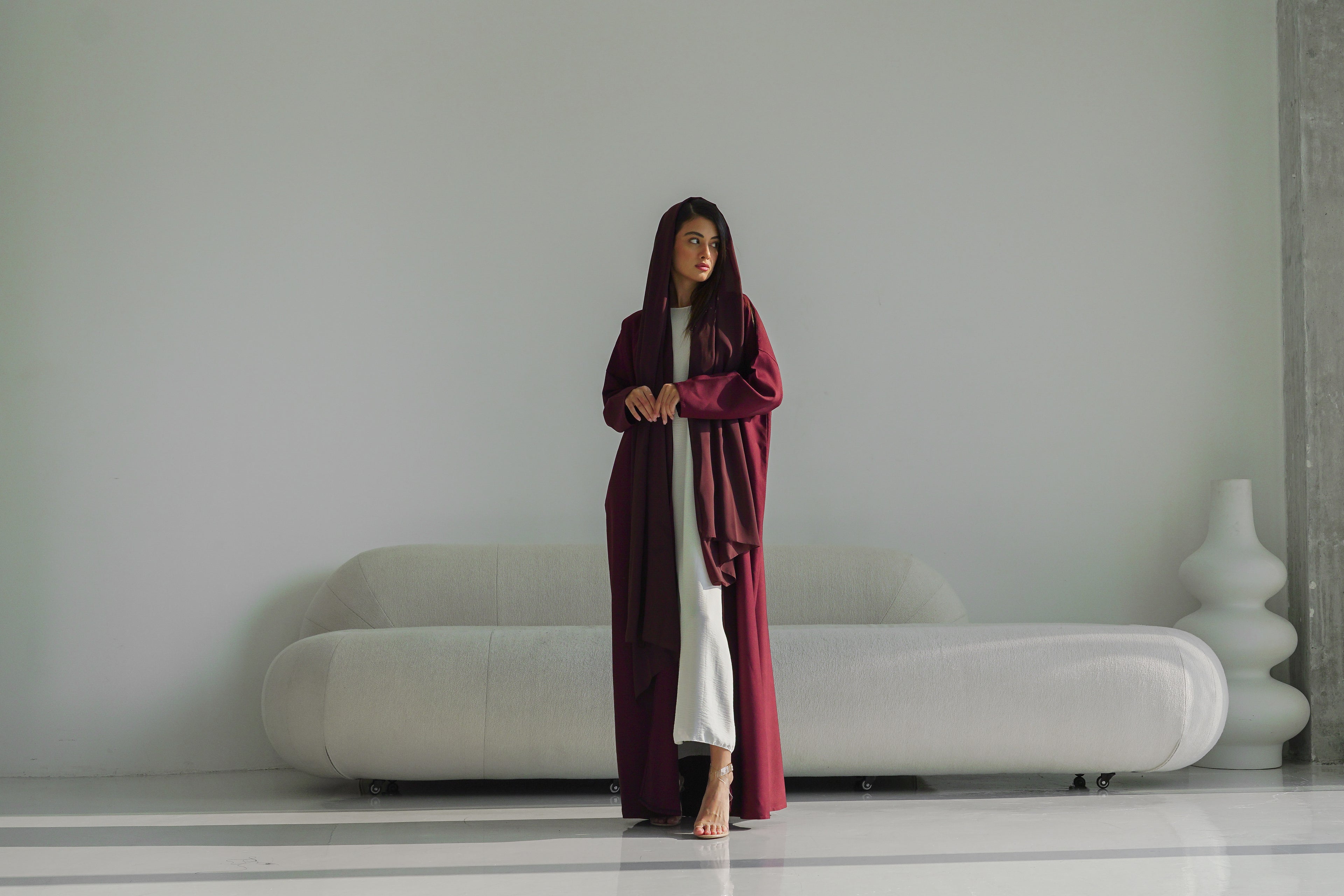 Woman in a long burgundy coat standing in a minimalistic room with a white sofa.