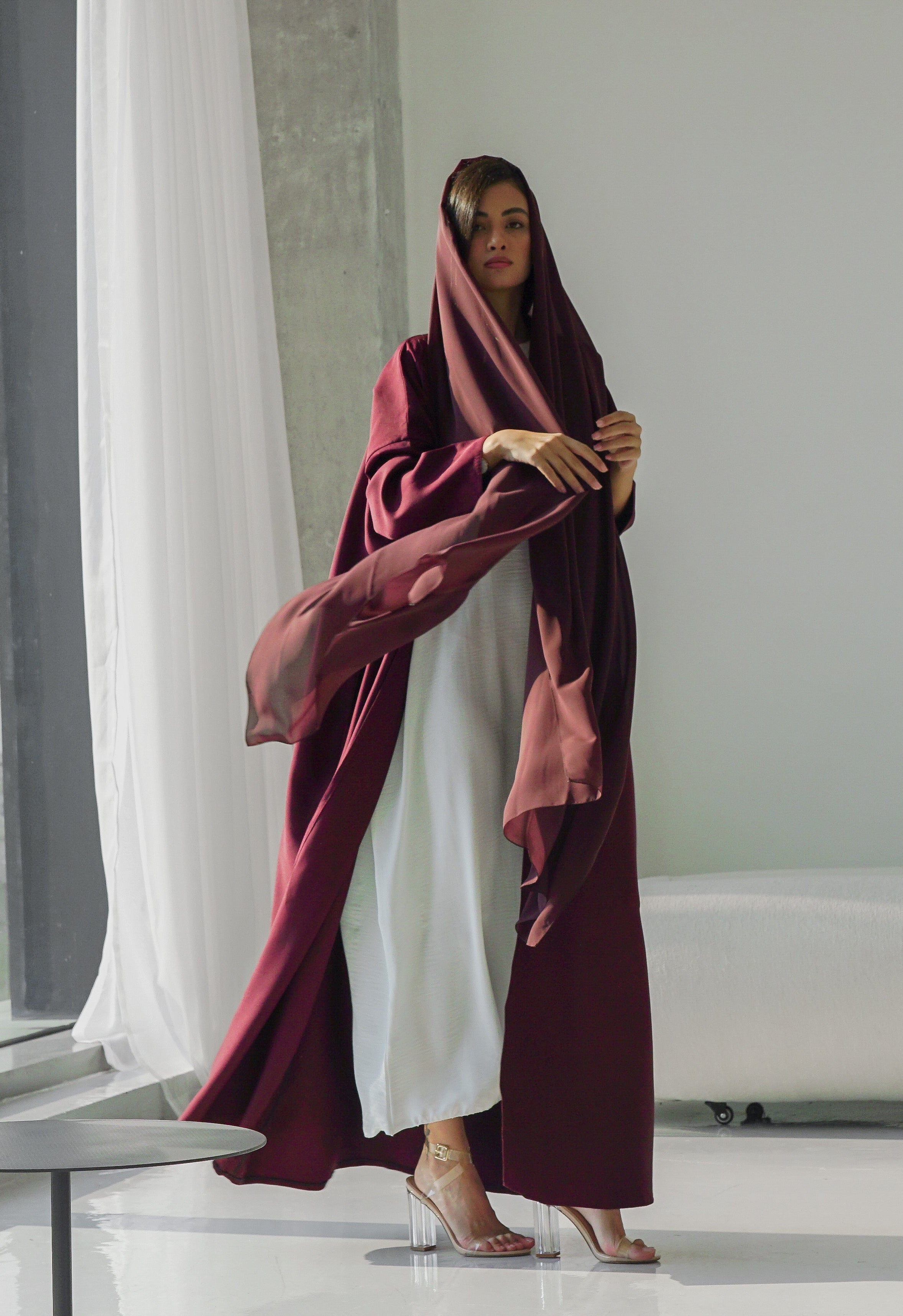 Woman in a burgundy abaya standing in a minimalistic room with a large mirror.