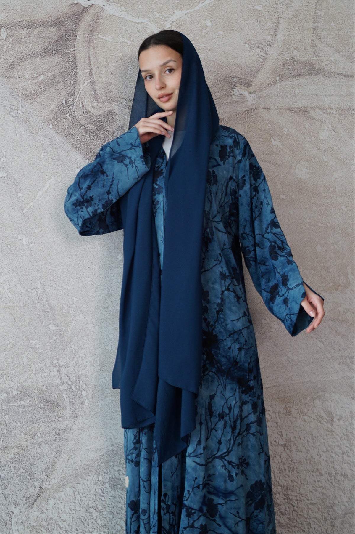 A woman wearing a long-sleeved navy blue abaya with a blue floral pattern and a solid navy blue headscarf, standing against a textured gray concrete wall.