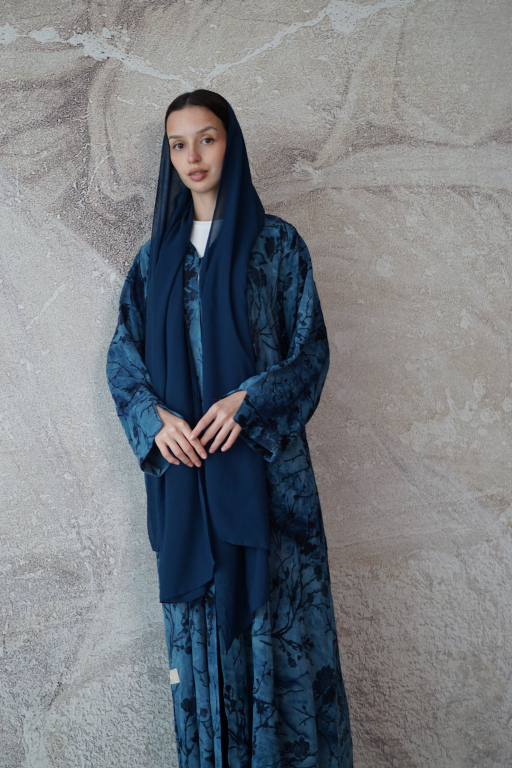 A woman wearing a dark blue patterned long robe with matching headscarf and trousers, standing against a textured gray wall.