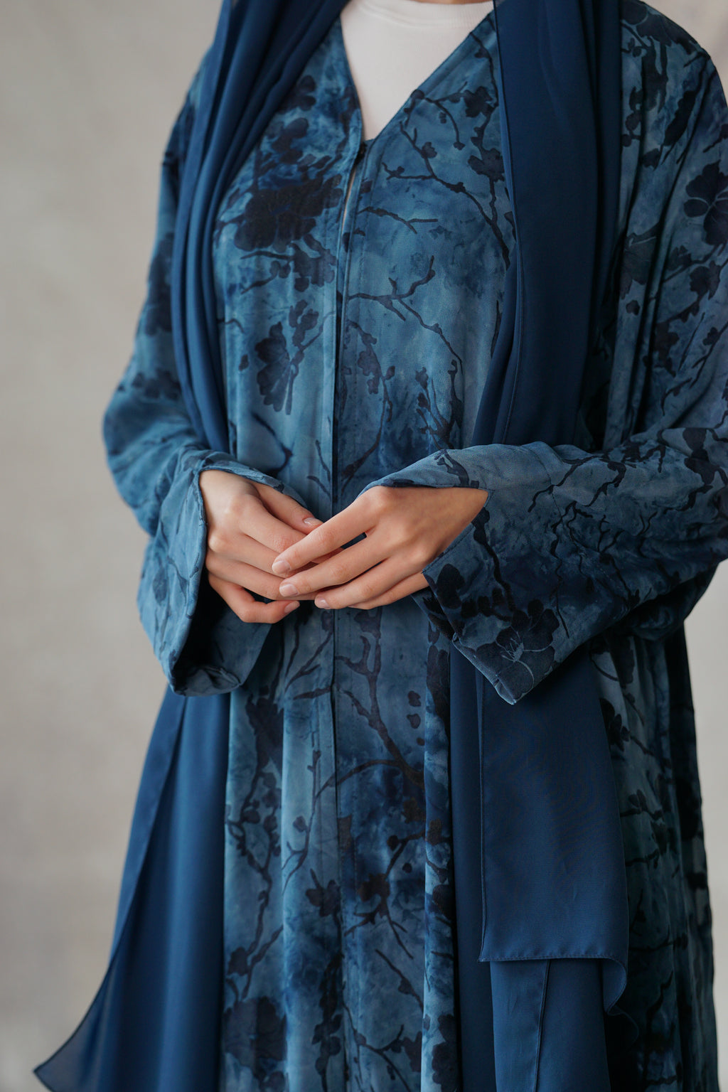 A person wearing a flowing blue robe with a dark floral or branch-like pattern, layered over a white top, against a soft neutral studio background.
