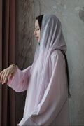 A woman wearing a light pink modest outfit with a matching headscarf, standing in profile against a textured beige wall.