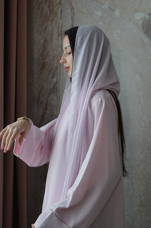 A woman wearing a light pink modest outfit with a matching headscarf, standing in profile against a textured beige wall.