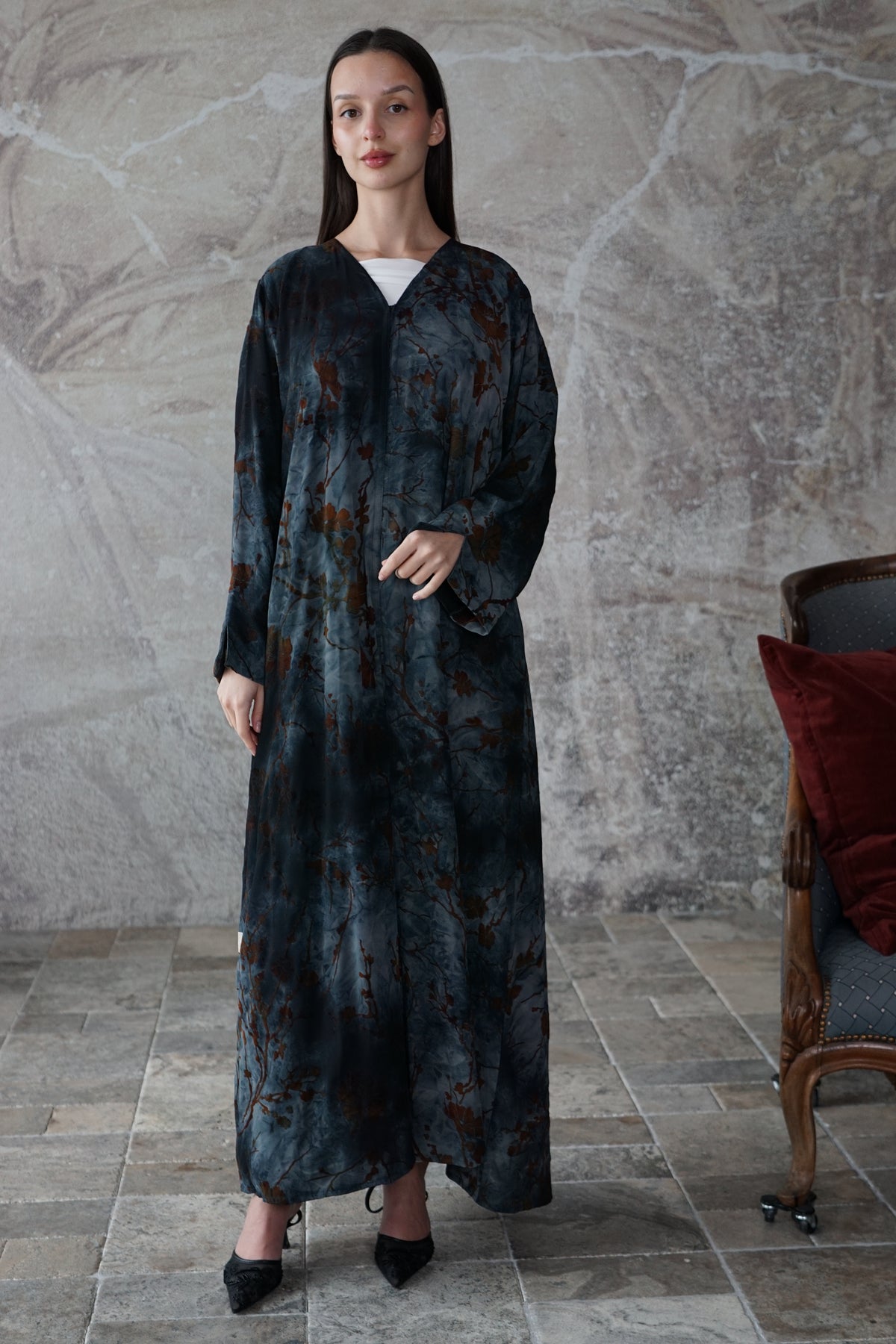 A woman stands indoors wearing a long, loose-fitting maxi dress with a dark blue and black abstract print and brown accents. The dress has a V-neck, long sleeves, and a front button closure.