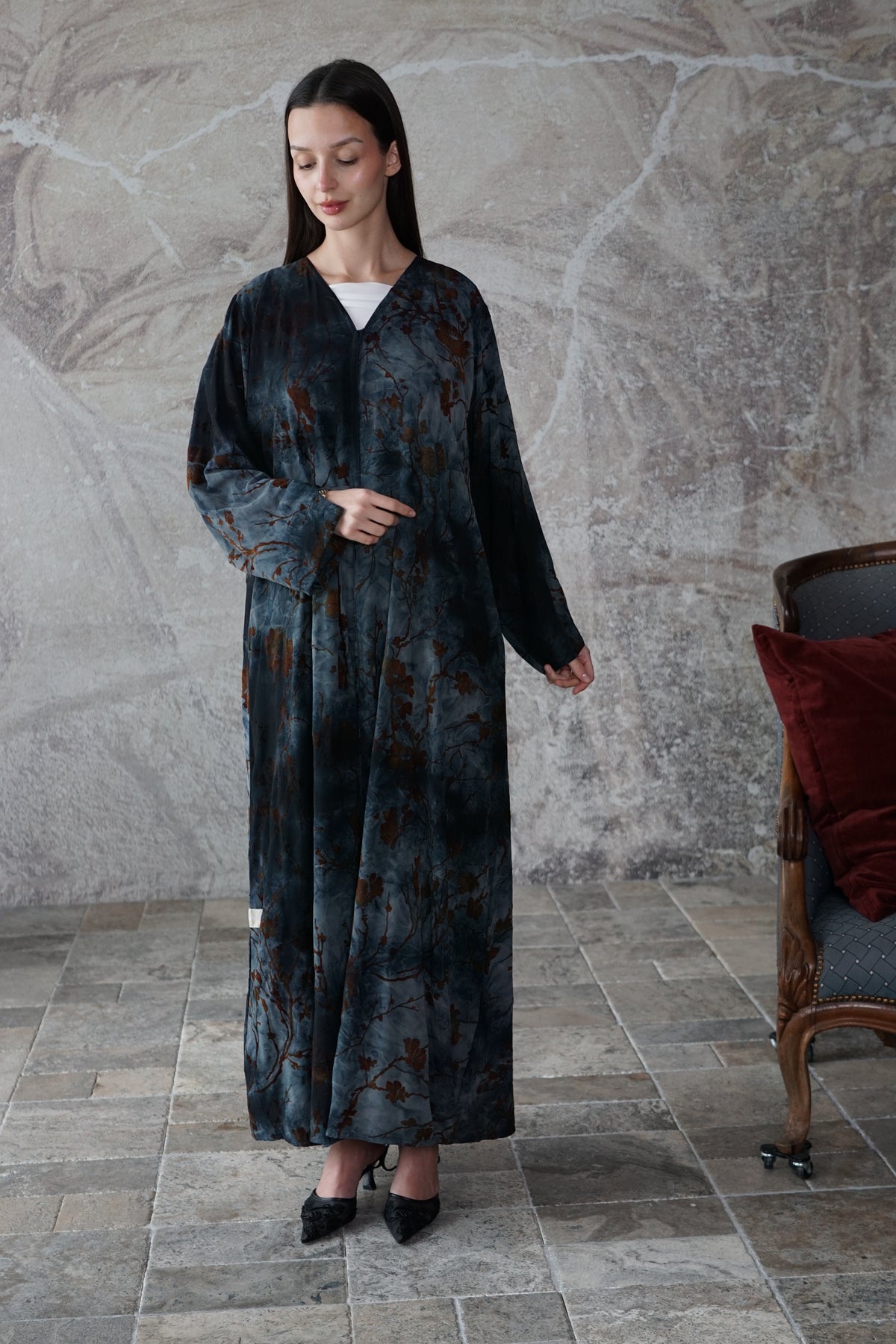 A woman stands indoors wearing a long dark blue-gray robe with an abstract brown and black marbled pattern. The robe has a V-neck, long sleeves, and a loose, flowing silhouette.