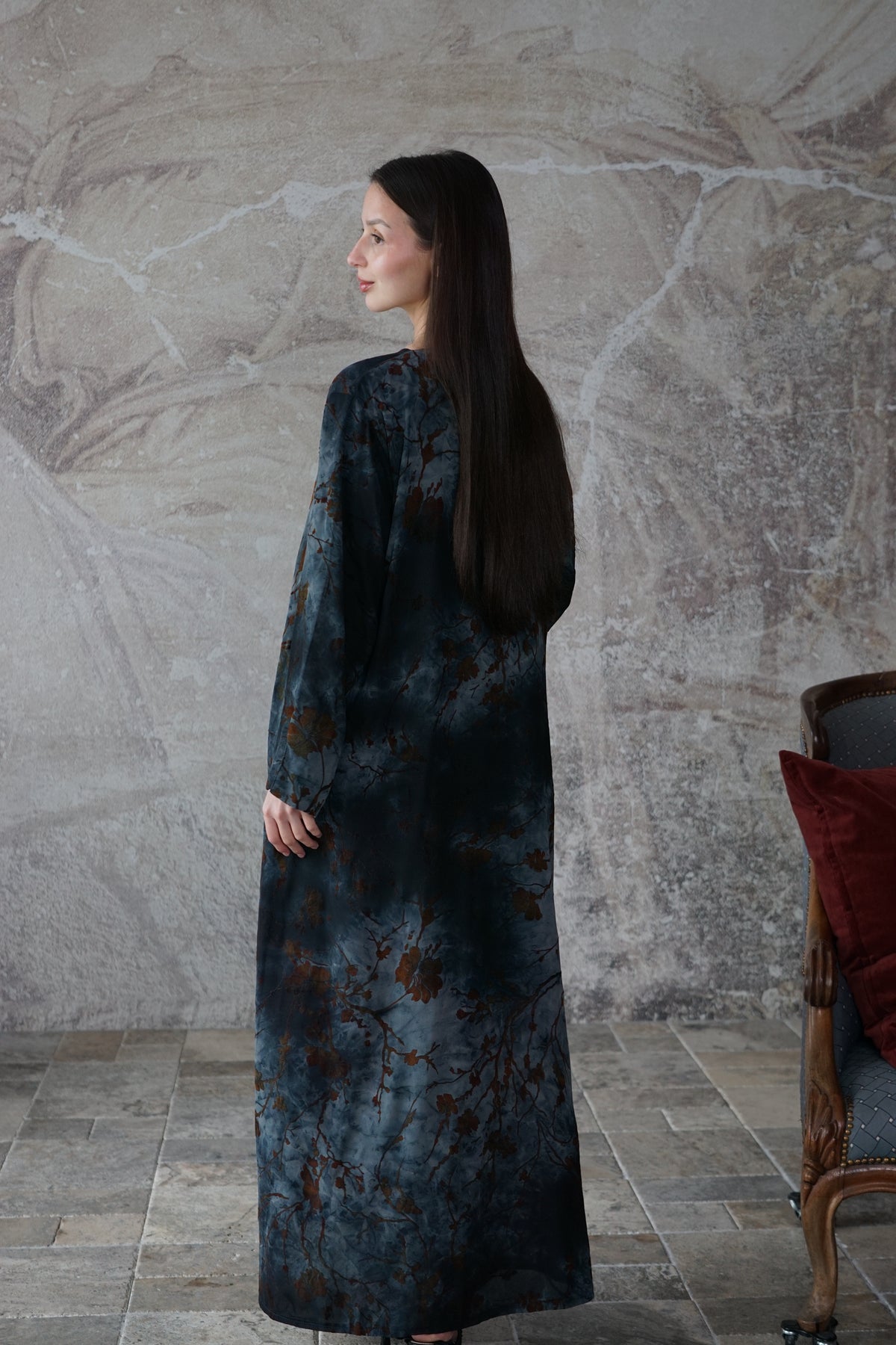 A woman with long dark hair is shown from the back wearing a long-sleeved, floor-length dress with a mottled blue and brown floral pattern, standing against a textured gray stone wall.