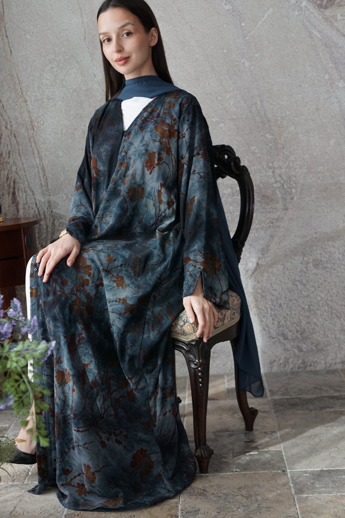 A woman seated on a dark wooden chair wearing a long-sleeved, loose-fitting kaftan-style dress in dark blue with an earthy brown floral pattern. The dress has a high collar and a draped front, styled in a rustic indoor setting with a textured gray wall and stone floor.