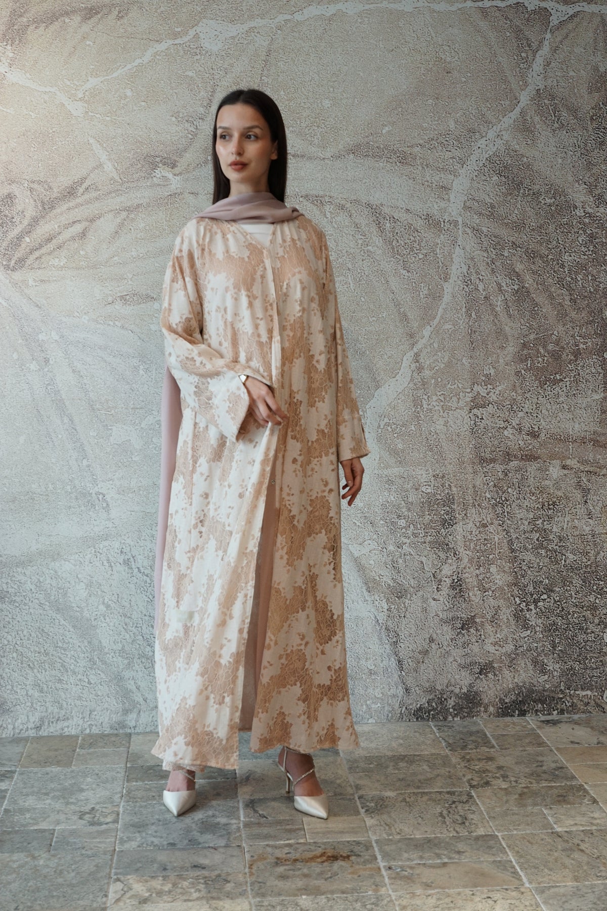 A woman models a long-sleeved, floor-length beige and white patterned abaya with a matching sheer dupatta, standing against a textured stone wall.